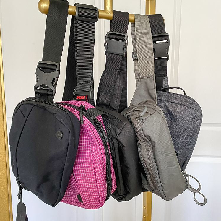 Five different concealed carry fanny packs hanging from a clothing rack. To black, two grey, and one pink.